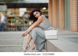 Woman sitting outdoors wearing a sleeveless striped jumpsuit and white handbag. | cheap clothes