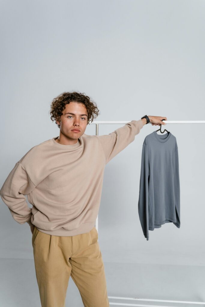 Young man with curly hair posing in a minimalist studio with a sweater on a hanger. | cheap clothes